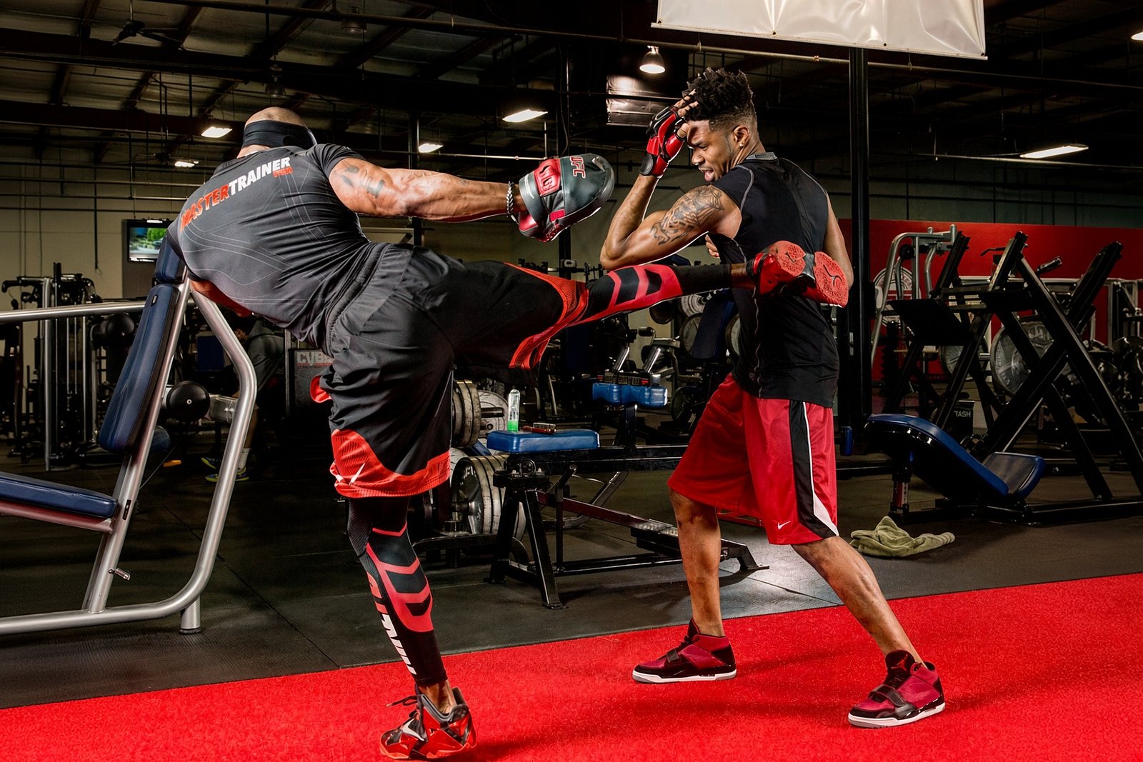 The author, in a Master Trainer shirt, throwing a high kick to a focus mitt held up by a fighter in red shorts on a red mat in a gym